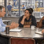 Painted factory workers and a manager in discussion beside the production floor.