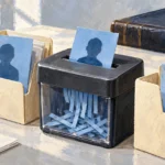 A painted paper shredder cuts a blue profile card while matching cards remain in two file folders beside a closed ledger.